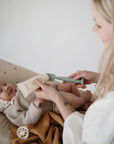 A smiling baby in a knit bonnet lies on a blanket as an adult uses the Mushie Diaper Cream Applicator to gently apply cream.