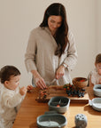 A woman slices fruit at a table with two young children, one eating and one holding a strawberry. mushie Lunchboxes with built-in compartments are on the table.