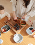 A woman cuts fruit on a wooden board while two young children sit at a table with mushie Lunchboxes featuring built-in compartments. Divided plates hold strawberries and blueberries, with extra berries and a wooden bowl on the table.