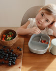 A young girl sits at a wooden table with strawberries, blueberries, and a mushie Lunchbox with built-in compartments for fruit. She rests her head on her hand and smiles gently.