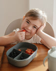 A young child smiles at the table, holding a strawberry by her mouth. In front of her is a mushie Lunchbox with compartments filled with strawberries and blueberries, and a light green sippy cup sits nearby.