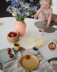 A young child in a light pink outfit reaches toward a white table set with mushie Round Dinnerware Plates, plastic cups, strawberries, a striped cloth, a pink vase of wildflowers, and a wicker basket.