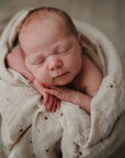 A newborn rests peacefully with eyes closed, swaddled in a mushie Organic Cotton Muslin Swaddle Blanket in beige with small star patterns; the softly blurred background adds to the calm scene.