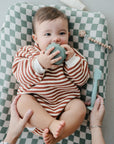 A baby in a red and white striped outfit lies on a mushie Extra Soft Muslin Changing Pad Cover in green and white checks, smiling and holding a teal cup, while an adult’s hands gently hold the baby’s legs. A pacifier clip is nearby.