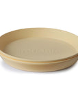The mushie Round Dinnerware Plates are stacked, round, beige plastic plates with "mushie" embossed in the center; these non-toxic plates are both stylish and dishwasher safe.