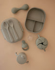A set of pale green, food-grade silicone baby feeding items by mushie on a beige surface, including a divided plate, bowl with lid, spoon, fork, sippy cup, snack cup, teether, pacifier, and the mushie Silicone Baby Rattle Toy for playful sensory fun.