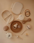 A flat lay of neutral-toned baby items—including the Mushie Classic Silicone Suction Plate—alongside cups, teething toys, a pacifier, toddler plates, a beaded bracelet, food feeder, and rattle on a beige background.