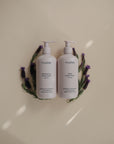 Two white pump bottles of mushie Lavender Baby Body Lotion sit side by side on a beige surface, accented by semicircular lavender sprigs to evoke a gentle lavender scent and natural ingredients.