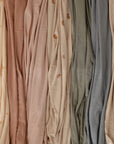 Mushie Stretchy Swaddle fabric swatches—cream with gold sun prints, dusty rose, beige with brown rainbows, sage green, and slate blue—are shown side by side in soft folds, ideal for a stretchy swaddle or breathable baby blanket.