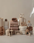 A neatly arranged display of mushie Lunchbox products in neutral tones, featuring non-toxic plastic containers, soft towels, pacifiers, teething rings, bottles, and folded blankets in beige, tan, and brown shades.