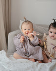 Two babies with small ponytails sit on a couch in neutral dresses, each holding pacifier clips. One baby smiles and chews on the mushie Flower Training Toothbrush, while the other gazes at the camera. A soft blanket covers the couch.