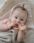 A light-skinned, blue-eyed baby with a tuft of hair tied up lies on the mushie Extra Soft Muslin Changing Pad Cover, wrapped in a beige towel, holding a teether and looking up with a dab of cream on their cheek.