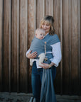 A woman smiles down at her baby in a grey wrap, who clutches the mushie Silicone Pacifier Clip | Luna—an adorable and practical accessory—while looking at the camera.