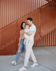 A smiling couple walks outdoors by a rust-colored staircase, holding a baby using the mushie Silicone Pacifier Clip | Luna. The woman wears a beige top and ripped jeans; the man is in a white shirt and pants. The baby is in light clothing.