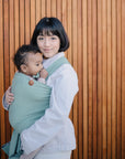 A woman with short dark hair holds a baby in a green carrier before a wooden slat wall. The baby’s mushie Silicone Pacifier Clip | Hera adds minimalist style, as both look calm and content.