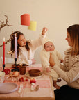 A woman holds a baby in white while a child playfully tugs the baby’s hat. On the decorated table are candles, fruit, and Mushie's Classic Silicone Suction Plate, adding to the warm, festive atmosphere.
