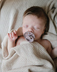 A sleeping baby with light brown hair lies peacefully on a soft beige blanket, holding a gray pacifier and partially wrapped in a mushie Organic Cotton Muslin Swaddle Blanket.