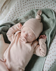 A newborn sleeps soundly on a mushie Extra Soft Muslin Changing Pad Cover, dressed in pink with a matching hat, wrapped in a green blanket, and soothingly sucking on a pacifier.
