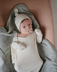 A baby in a cream hat and onesie lies on the mushie Extra Soft Muslin Changing Pad Cover, wrapped in a gray blanket and sucking a pacifier while looking up.