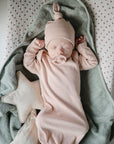 A newborn wearing a pink hat and gown sleeps on a mushie Extra Soft Muslin Changing Pad Cover, holding a pacifier. A soft star-shaped pillow rests beside them, with the baby’s arms raised near their head.