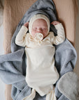 A newborn in a cream outfit and hat sleeps on a soft blue-gray blanket, holding a pacifier, atop the mushie Extra Soft Muslin Changing Pad Cover with a peach quilt underneath.