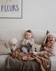 Two babies sit on a couch with a small white dog. One laughs in a bonnet, while the other holds natural wood toys. A brown blanket covers part of the couch, and above hangs a FLEURS poster. Nearby is the mushie Silicone Pacifier Clip | Mila.