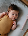 A baby in a mustard yellow sleeveless outfit lies on a mushie Extra Soft Muslin Changing Pad Cover next to a pink silicone spatula.