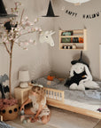 A cozy, decorated children’s bedroom features Halloween accents—including a Happy Halloween banner and bat decor—plus a witch-hat-wearing stuffed bunny and a child playing on the floor with the mushie Phone Press Toy for sensory exploration and fine motor skill building.