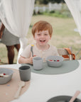A young red-haired child smiles at an outdoor table holding a fork, surrounded by bowls and mushie Dinnerware Cups. Colorful placemats decorate the table, while sheer white curtains and greenery create a gentle backdrop.