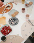 A meal table set with mushie Silicone Placemats, pastel plates and cups, cutlery, snacks, a glass water bottle, and a silicone bib. Children are seated around it enjoying raspberries, blueberries, crackers, and more.