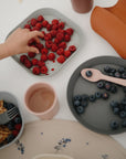 A child's hand reaches for raspberries on a gray mushie Round Dinnerware Plate, surrounded by dishwasher-safe plates of blueberries, a cup, a silicone bib, and small snacks on a white table.