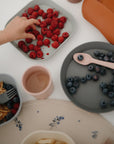 A child's hand reaches for raspberries on a plate, surrounded by blueberries, pancakes, and a cup—each set on a food-grade silicone placemat by mushie atop a white table.