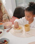 Two young children sit at a table set with mushie Silicone Placemats. One eats chips; the other, holding a stuffed toy, looks down. Their bowls of cereal and fruit rest on these colorful, food-grade mats ready for mealtime fun.