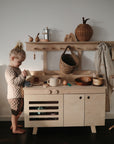 A young child with blonde hair in a bun plays at a light wood play kitchen, surrounded by baskets, cups, bowls, and a mushie Organic Cotton Muslin Swaddle Blanket in a cozy, minimalist room.