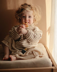 A young child with curly blonde hair sits on a mushie Extra Soft Muslin Changing Pad Cover by a sunlit window, wearing a striped beige outfit and holding a yellow teething toy, with a pacifier resting nearby.