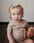 A light-skinned young child with curly reddish hair and blue eyes, wearing a beige floral onesie and headband, sits indoors holding a toy and the mushie Silicone Pacifier Clip | Hera, gazing curiously at the camera.
