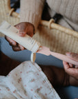 A person squeezes lotion onto the Mushie Diaper Cream Applicator, preparing to apply it to a baby in a white floral outfit. The pink silicone applicator and partially visible baby are in the foreground.