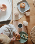 Two young children sit at a wooden table with woven placemats, enjoying pastries and bananas served on mushie Square Dinnerware Plates. The pastel BPA-free plates create a cozy, neatly arranged scene viewed from above.