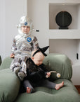 Two young children sit on a green couch—one in a shiny astronaut costume, the other in a sparkly witch dress holding a FRIGG Daisy Natural Rubber Pacifier by FRIGG and a beaded toy. Decor items are displayed on shelves in the background.