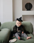 A baby in a black sparkly witch costume and pointed hat, using the mushie Silicone Pacifier Clip | Hera, sits on a green couch near a paint palette, with white shelves and round decor enhancing the room’s minimalist style.