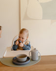 A baby in a high chair holds a teething toy, wearing a blue jacket and bib. An adult woman leans in from the left, while Mushie’s Classic Silicone Suction Plate sits on the table. A beige abstract painting hangs on the wall behind them.