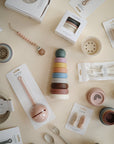 A flat lay of mushie baby feeding items, non-toxic teething toys, and the mushie Stacking Rings Toy arranged on a cream surface, including spoons, bowls, forks, silicone baby toys, and branded packaging.