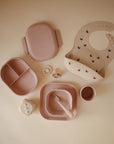 The mushie Lunchbox set features pastel pink and beige silicone baby feeding items, including a compartment plate, bowl, cup, butterfly bib, fork, spoon, teether, pacifier, and dice-shaped toy—all displayed on a beige surface.