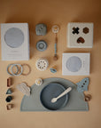 A flat lay of baby items in soft neutrals, featuring the Mushie Classic Silicone Suction Plate with spoon, stacking toys, toddler plates, teething rings, a shape sorter cube, two boxed products, and cute teethers.