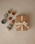 A mushie Stacking Rings Toy—pastel-colored, non-toxic rings—sits neatly arranged beside a brown gift box tied with a white ribbon on a beige surface.