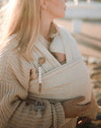 A blonde woman in a knitted sweater holds a baby wrapped in a soft beige sling, with a beaded cross clip and a FRIGG Andersen Fairytale Natural Rubber Pacifier from FRIGG attached. Warm sunlight fills the outdoor scene.
