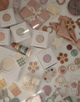 A flat lay of mushie’s Flower Training Toothbrush, pastel baby toys, teethers, stacking rings, cloths, and gift boxes arranged neatly on a white surface in soft pink, beige, green, and yellow tones.