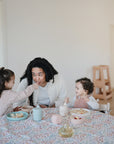A woman sits at a table with two children in a bright room. One child feeds her from a Mushie Classic Silicone Suction Plate, while the other smiles. Bowls, cups, and a patterned tablecloth are set on the table.
