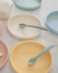 Colorful, non-toxic Mushie Classic Silicone Suction Plates in pastel yellow, pink, blue, green, and cream with matching spoons, a sippy cup, and a pink textured utensil holder arranged on white marble—perfect for toddlers.