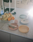 A marble kitchen counter displays a fruit bowl with bananas and oranges, three Mushie Classic Silicone Suction Plates with matching spoons, four white candles, and a bright light shining in the background.